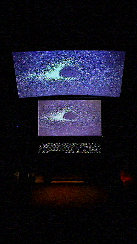 Two screen workstation with a Dell P2214H monitor on the desk and a larger curved Samsung ViewFinity S65UA monitor mounted above, both showing the same grainy blue and purple edit of the Jean-Pierre Luminet black hole image; Logitech MX Keys S Keyboard backlight is visible; dark, moody room with low ambient lighting; tone is contemplative and cinematic